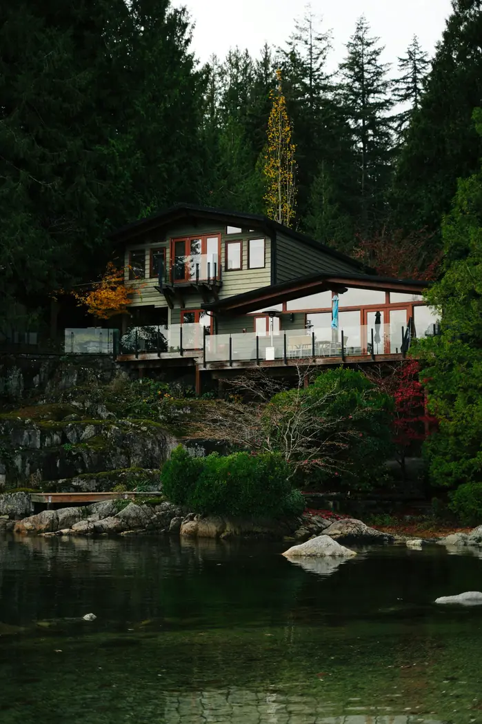 Mid-century modern waterfront home with glass walls and a low-slope roof, perched above a rocky lake shoreline among evergreens